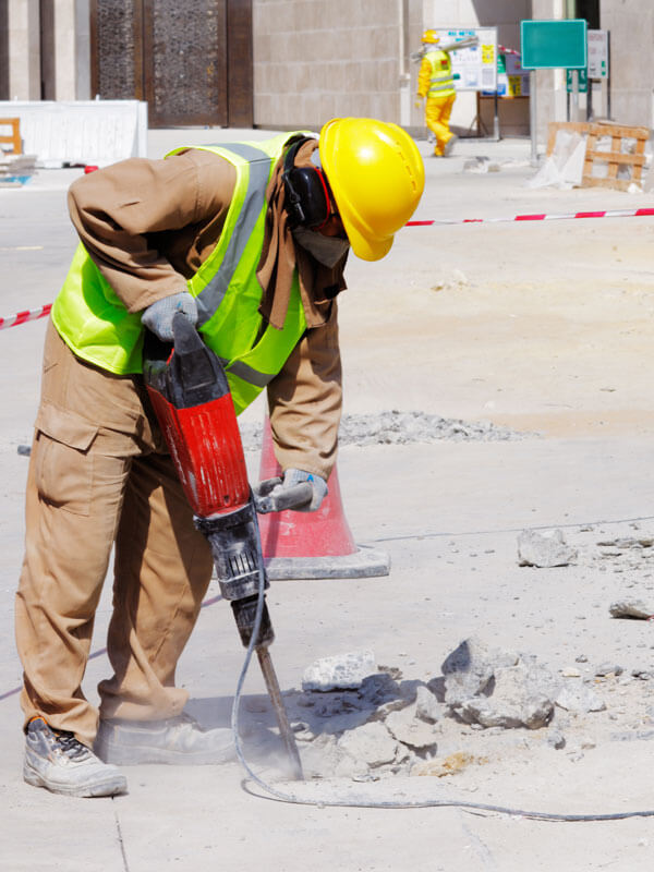 Workman using an automatic drill in the ground, wearing protective ear defenders and hard hat